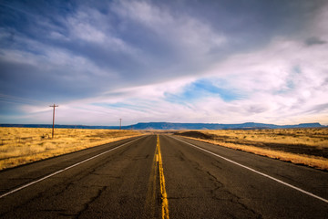 A highway cutting between golden pasture land with mountains in the distance under a cloudy sky in a Colorado countryside landscape