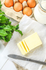 A bar of butter is cut into pieces on a wooden board with a knife, surrounded by milk, eggs and parsley on a white table. Ingredients for cooking.