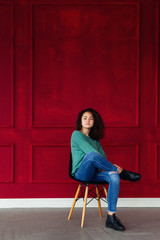 brunette with curly hair sitting on a chair on a background of a decorated red wall