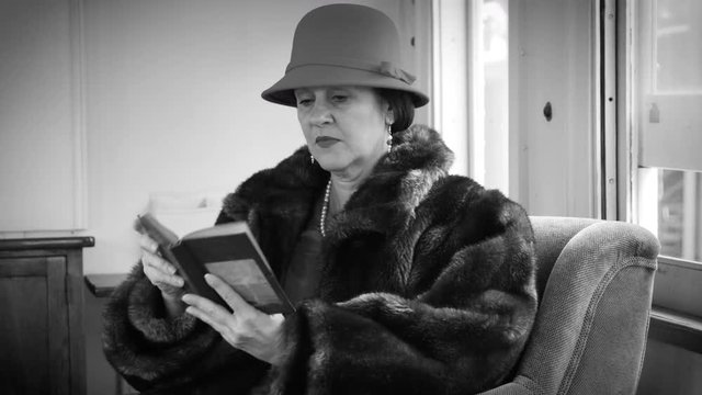 A lovely passenger train traveler in stylish 1930s or 1940s period attire sitting in the lounge car of a vintage railcar quietly reading a book during a scheduled stop along her journey.
