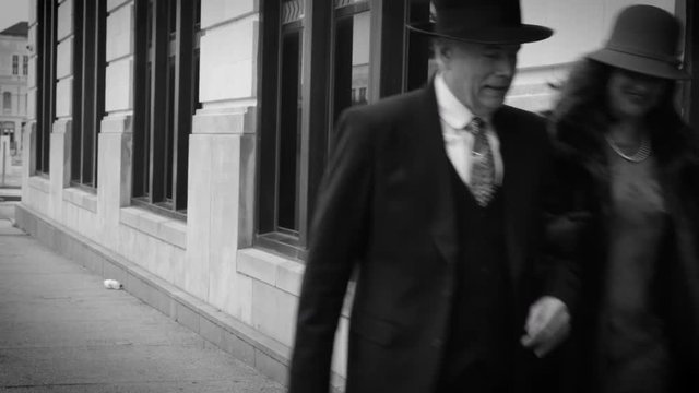 Period piece footage of a lady and a gentleman walking past enormous windows of historic building with the woman wearing fur coat and carrying vintage style travel bag and man sporting a fedora.