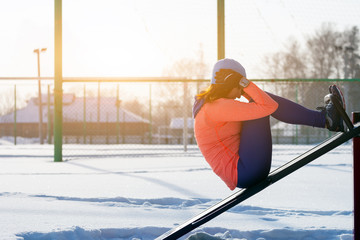 A young woman in a bright blue hat, orange sweatshirt and elk skewers presses a press on a vertical simulator on a sports field on a bright winter day