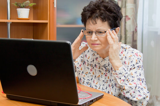 Cute serious senior woman holding eyeglasses and working with the notebook at home 