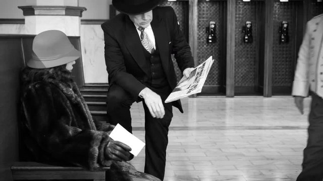Period piece of lady in fur coat and round bowler cap and a gentleman sporting a fedora who are in a large lobby talking about newspaper article when porter or bellhop comes to assist with luggage.