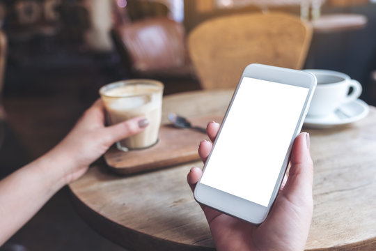 Mockup image of hands holding white mobile phone with blank desktop screen and a glass of coffee on wooden table in cafe