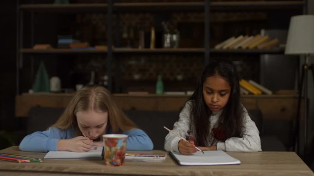 Charming little multiracial preteen girls painting using watercolors and gouache while sitting at desk in domestic room. Creative children drawing a picture with paintbrushes and watercolors at home.