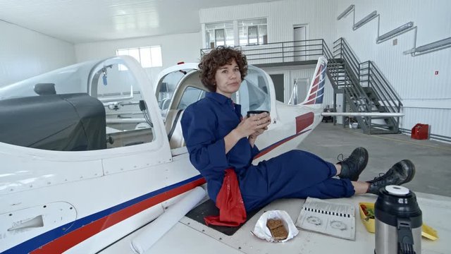 Tilt down of female aircraft mechanic sitting on wing of light jet airplane in hangar and drinking tea from thermos cup while having lunch