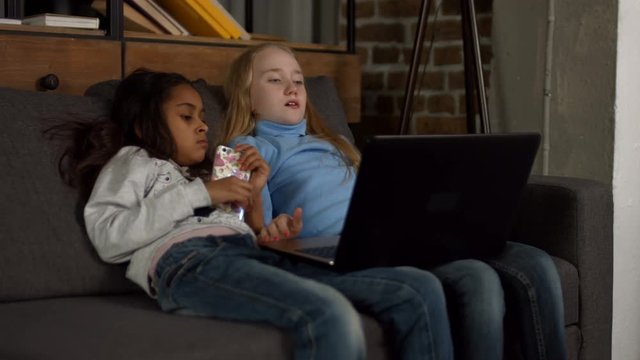 Diverse positive little girlfriends watching on line tv show on laptop while spending leisure together at home. Relaxed preteen multiracial kds sitting on the couch in domestic room using lapyop pc.
