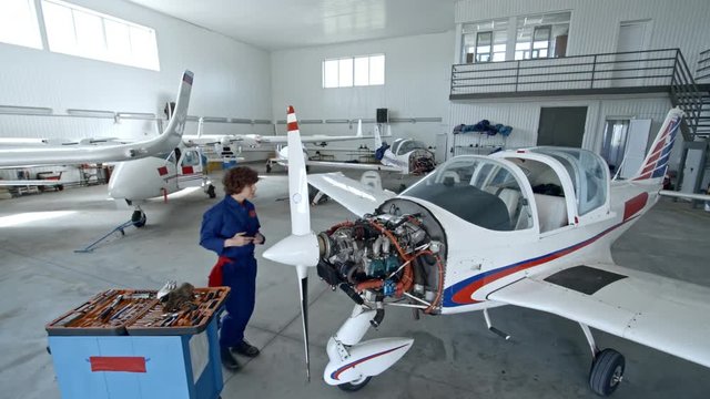 Tilt down high angle shot of female technician in uniform walking to cart with instruments, taking tool and fixing airplane engine in hangar