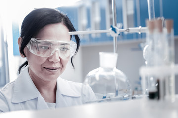 Monitoring the process. Mature brunette lady wearing safety glasses looking at a laboratory glassware fixed on a stand while conducting a chemical experiment.