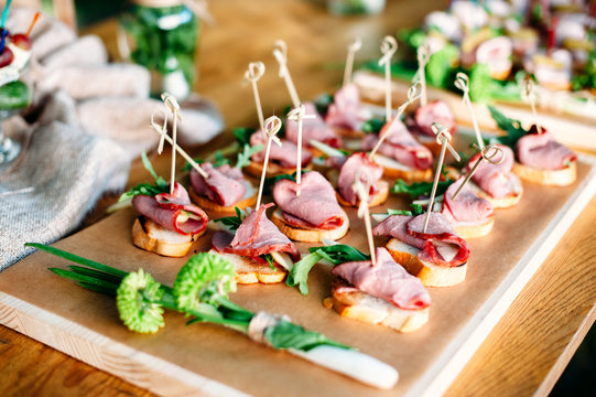Delicious catering banquet buffet table decorated in rustic style in the garden. Different snacks, sandwiches with ham and greenery on a wooden plate. Outdoor.