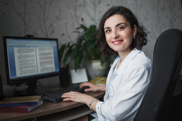 Smiling woman doctor in a with dark curly hair in white medical robe sitting at a table. On the table books, a computer monitor and a green plant. The horizontal frame.