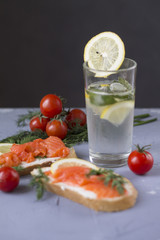 sandwich with red fish trout a piece of lemon and delicate cheese a glass of water with lemon mint and ice juicy red cherry tomatoes on a stone table