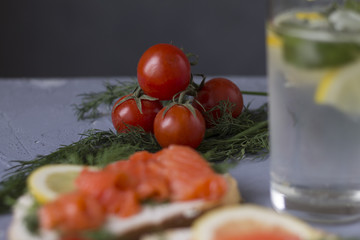 sandwich with red fish trout a piece of lemon and delicate cheese a glass of water with lemon mint and ice juicy red cherry tomatoes on a stone table