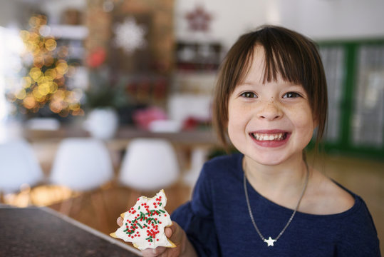 Smiling Girl in an open plan living room with an illuminated Christmas tree holding a homemade Christmas cookie with icing