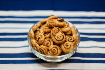 A horizontal lifestyle image of a glass bowl filled with homemade cookies, made of puff pastry, with sugar and cinnamon. On a surface, covered with blue and white stripy towel. Selective focus.