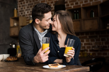 smiling couple with orange juice going to kiss at kitchen