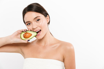 Beauty portrait of brunette asian woman biting and eating half of fresh ripe avocado, isolated over white background