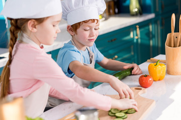 cute kids in chef hats and aprons preparing vegetable salad together in kitchen