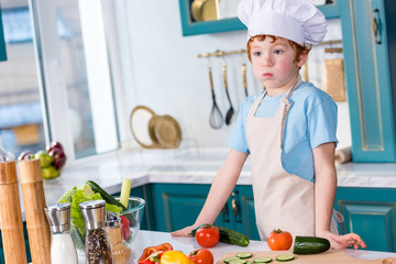 cute little boy in chef hat and apron looking away while cooking in kitchen