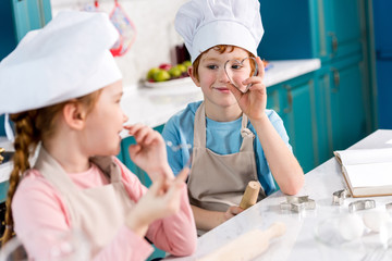 adorable children in chef hats and aprons smiling each other while cooking together in kitchen