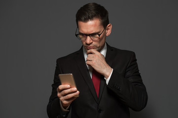 Serious thoughtful businessman with glasses and formal suit touching his face by hand and looking at the smartphone screen on gray background