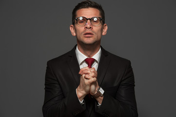 Portrait of middle-aged businessman in formal suit and glasses with crossed hand near his chest and praying against gray background