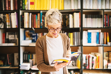 Portrait view of pretty cheerful female with glasses reading pages while standing in the library.