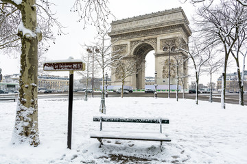 Winter in Paris in the snow. The Arc de Triomphe with a public bench covered in snow in the foreground. - obrazy, fototapety, plakaty