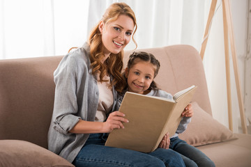 happy mother and daughter reading book and smiling at camera together at home