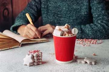Crédence de cuisine en verre imprimé Chocolat Tasse en papier de chocolat chaud avec de la guimauve devant des mains féminines écrivant quelque chose dans un ordinateur portable
