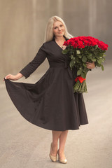 Portrait of beautiful blonde woman in black retro dress holding big bouquet of one hundred red roses outdoor