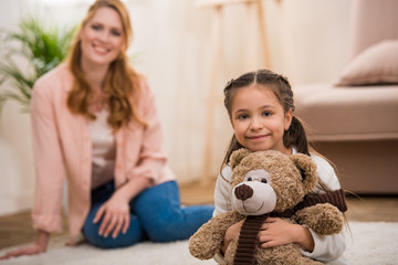 adorable little child holding teddy bear and smiling at camera while mother sitting behind at home
