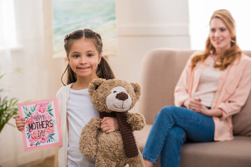 adorable child holding teddy bear and happy mothers day greeting card while mother sitting behind
