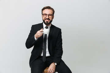 Joyous adult man wearing business suit and eyeglasses drinking coffee from cup and sitting on chair in office, isolated over white background