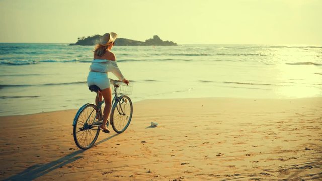 Woman in white and hat mooving on her bicycle along beach sand at summer time under burning sunset sky, lifestyle carefree.