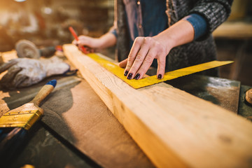 Close up view of hardworking focused professional serious carpenter woman holding ruler and pencil while making marks on the wood at the table in the fabric workshop.