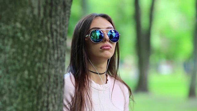 Young woman stands near tree in city park listens to music and dances. Beautiful girl in sunglasses with earpieces listens to music on the nature
