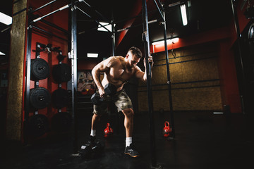 Powerful motivated bodybuilder shirtless guy holding dumbbell and doing triceps exercise while with another hand holding a vertical bar in the dark gym.