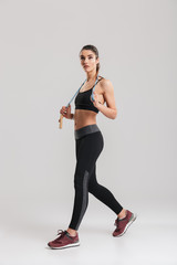 Full-length picture of slim woman looking aside and holding skipping rope on her neck, isolated over gray background