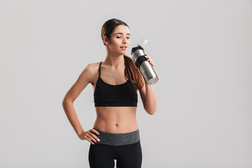Beautiful young fitness instructor wearing tracksuit looking aside and drinking fresh still water from metal bottle, isolated over gray background