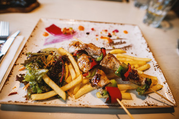 Close up focus view of fried skewers on a wooden stick and chips with salad on the plate in the restaurant.
