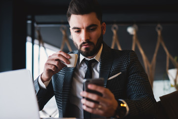 Attractive bearded suited man texting and drinking coffee while sitting in a restaurant.