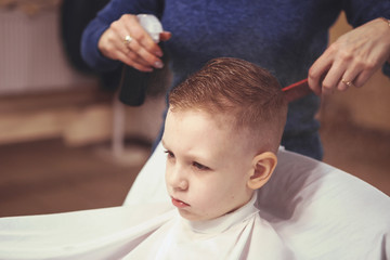 Fototapeta premium Little boy at the hairdresser. Child is scared of haircuts. Hairdresser's hands making hairstyle to little boy, close up