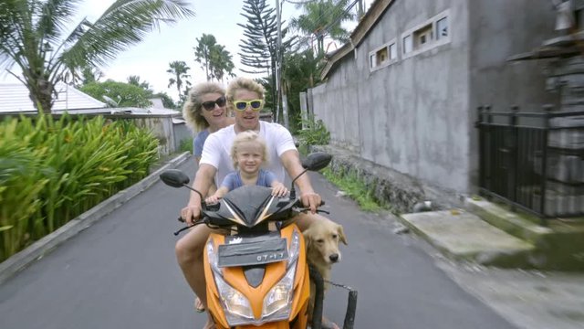 Excited family riding on scooter with dog in Bali: little boy and dad shouting and woman outstretching her arms