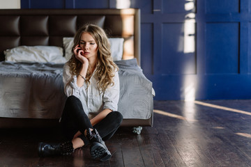 Obraz na płótnie Canvas A girl with long blond hair dressed in black jeans, a striped shirt and black shoes sitting on the bed with blue linens. Fashionable casual outfit.