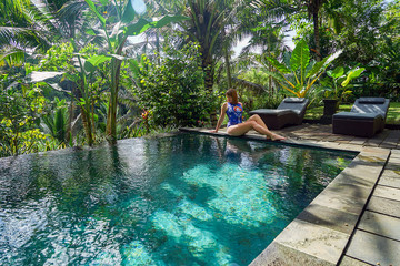 A young girl sits on the edge of an infinity pool in the jungle. Taken on the island of Bali in...