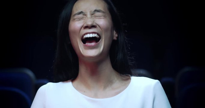 Slow motion reaction of an asian woman laughing while watching a funny movie at the movie theatre.
