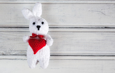 White toy Bunny holding a red heart in his paws on white wooden background
