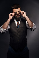 Obraz premium Low key portrait of serious man with dark hair, mustache and beard in white shirt, brown vest and colorful tie takes off round sunglasses
